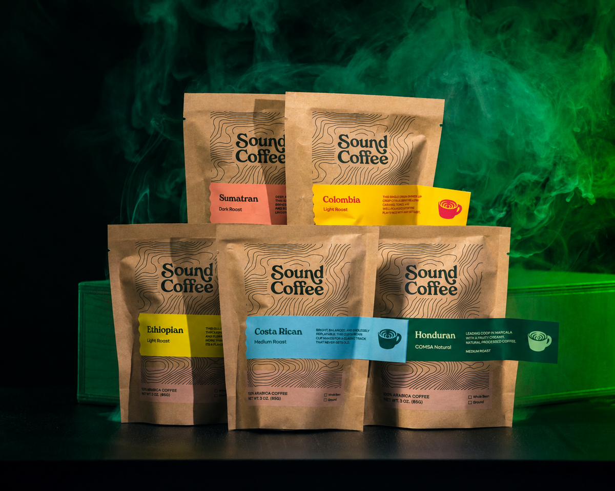 Five Sound Coffee branded coffee pouches arranged in two rows on a dark surface, with a green-lit smoky background. The front row includes Enterprise, Costa Rican, and Honduras coffees, and the back row includes Sumatra and Colombia coffees.