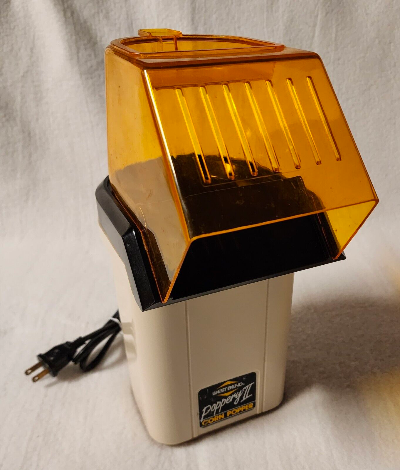 A vintage popcorn machine with a translucent amber-yellow lid, a cream-colored body, and a black top section, shown with its power cord on a white fabric background.