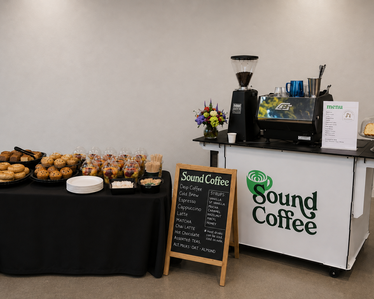 A Sound Coffee station setup with a white cart branded Sound Coffee, a black espresso machine, coffee grinder, and a menu board, alongside a table of pastries and coffee on a black cloth.