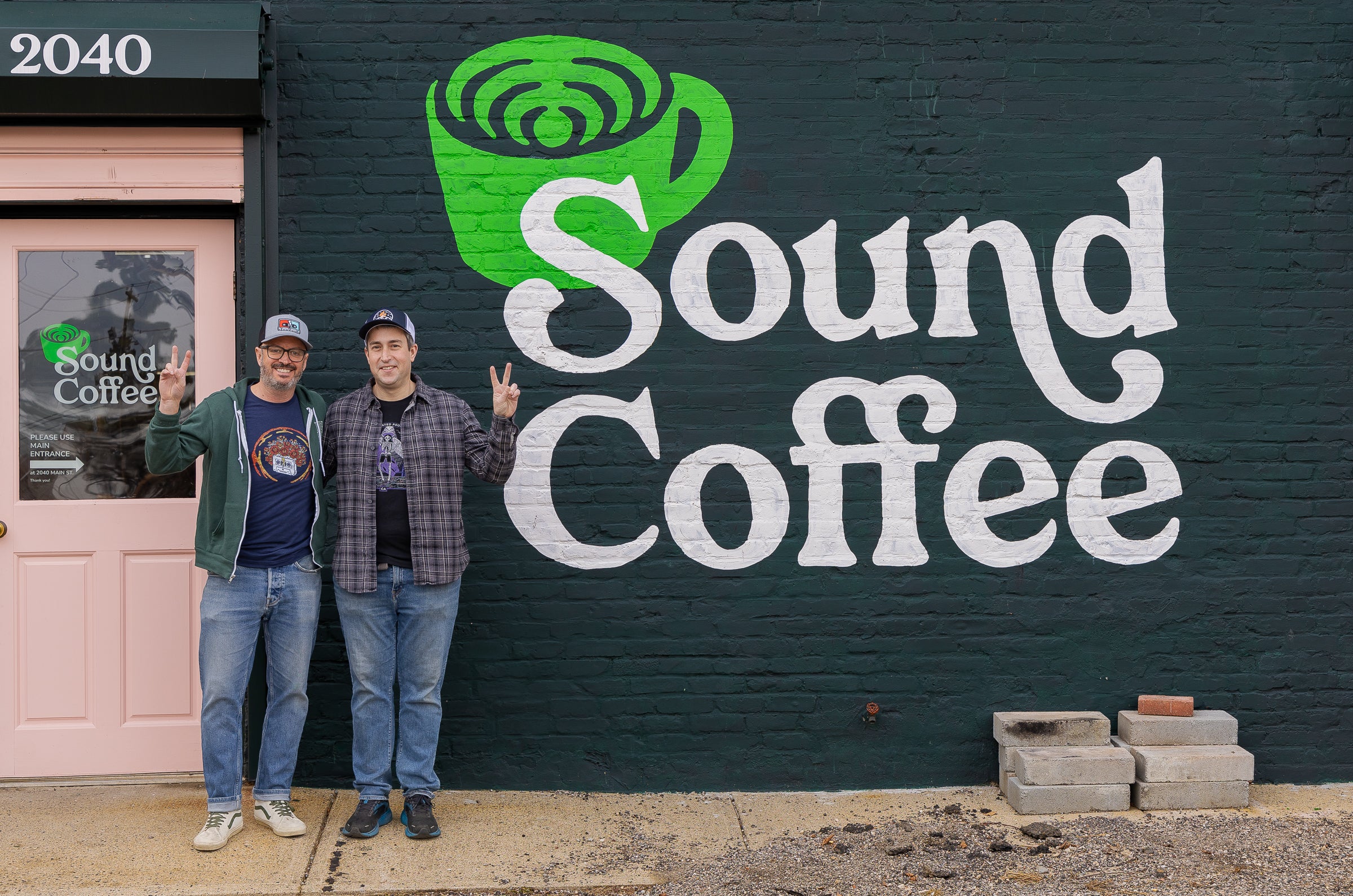 Two men standing in front of a dark painted brick wall with a large white 'Sound Coffee' sign and a green coffee cup logo. A beige door with 'Sound Coffee' in the window is visible on the left.