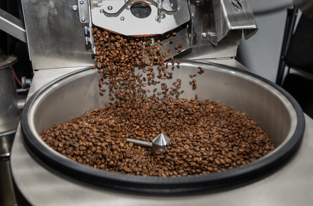 Roasted coffee beans falling from a metal hopper into a large stainless steel drum inside a coffee roaster.
