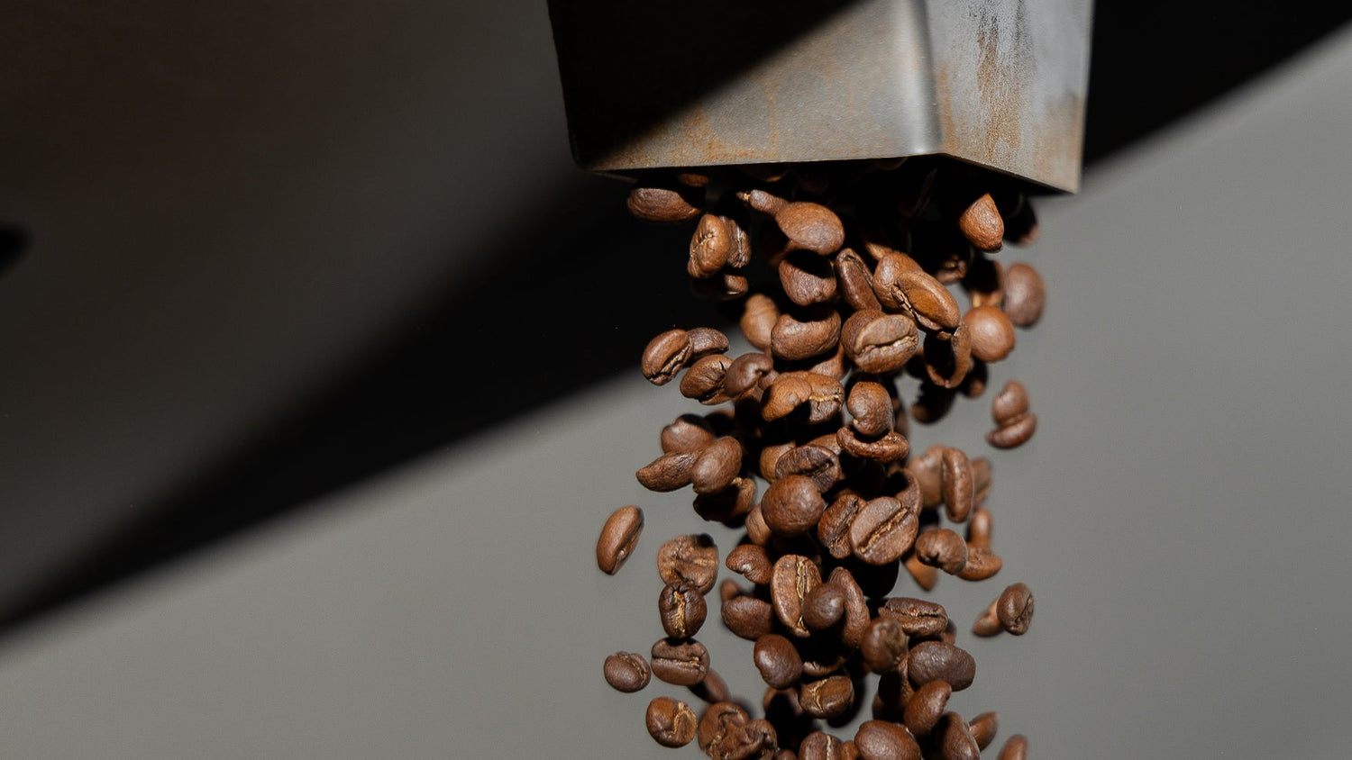 Roasted coffee beans falling from a metal hopper against a dark gray background.