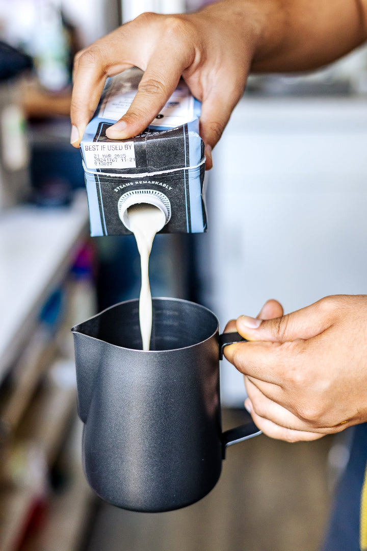 A close-up of a person pouring milk from a carton into a matte black milk pitcher, with the person's hands and arms visible against a softly blurred kitchen background.