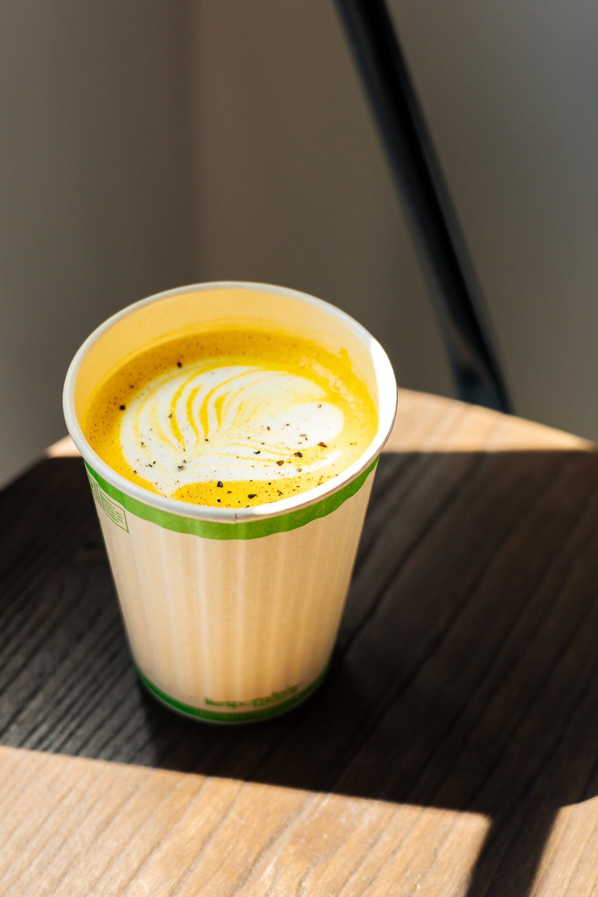 A paper coffee cup with a green stripe, filled with a yellow-orange latte and topped with white foam, sitting on a wooden table in natural light.