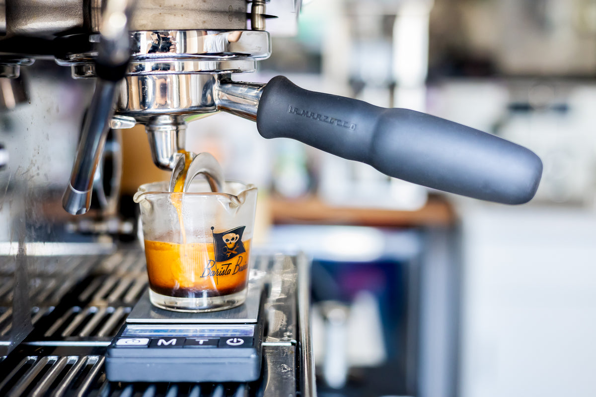 A close-up of an espresso machine dispensing espresso into a small glass measuring cup on a scale, with a blurred shop background.