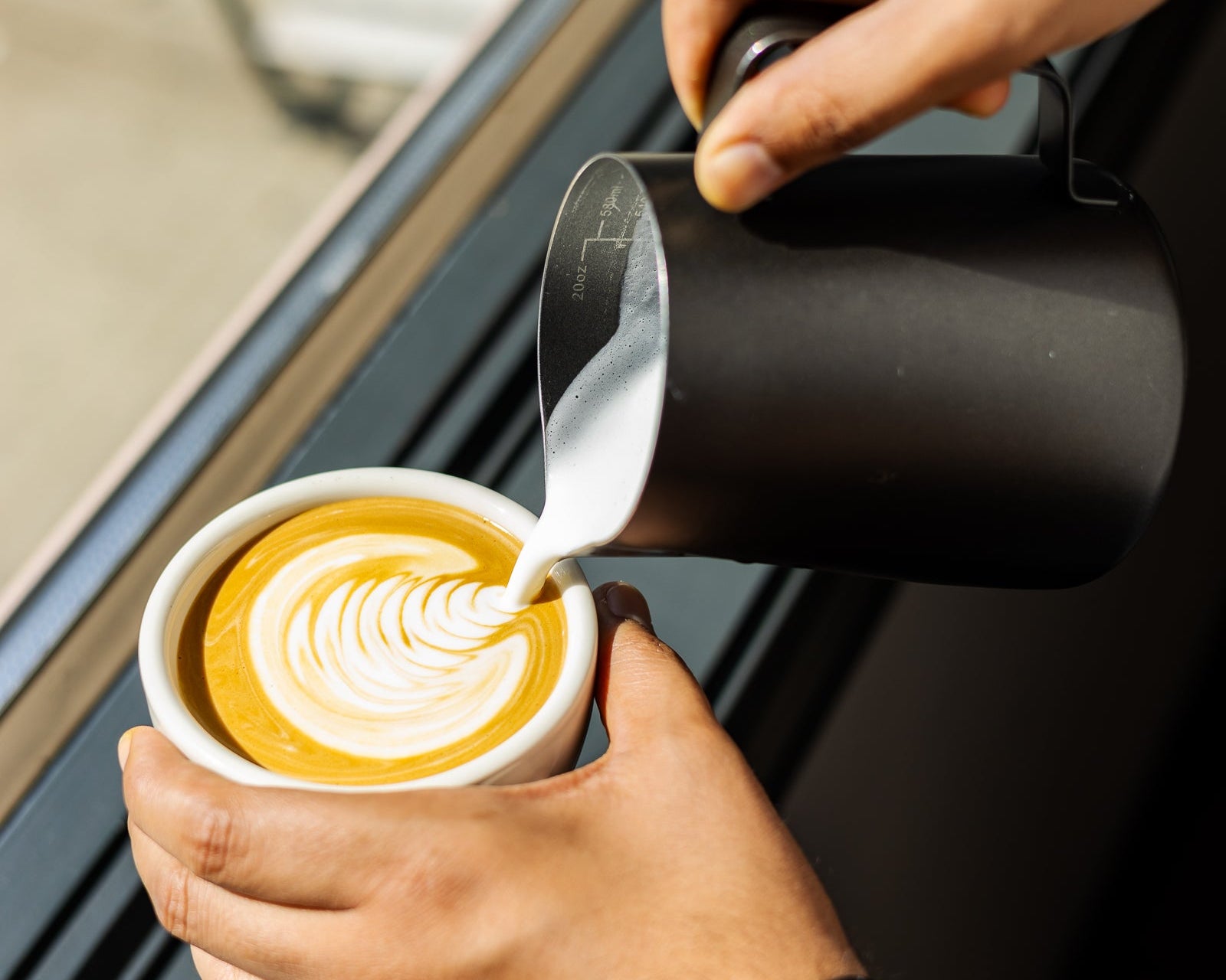 A close-up of a person pouring steamed milk from a matte black pitcher into a white cup of coffee with latte art, set near a window.