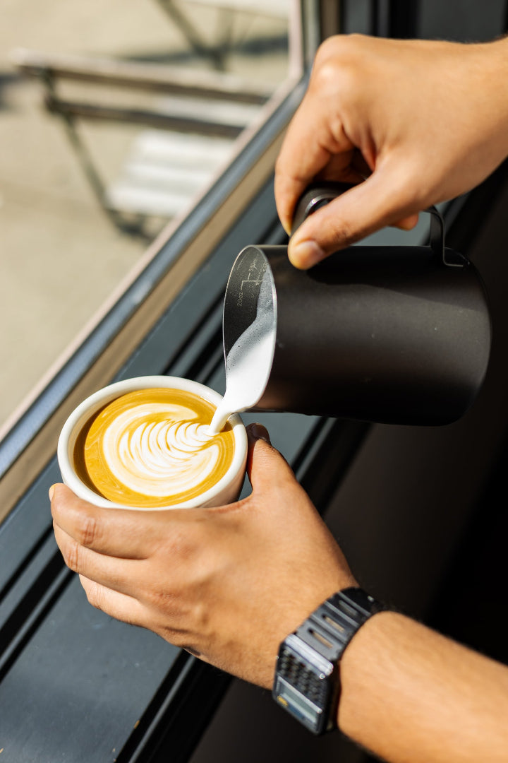 A close-up of a person pouring steamed milk from a matte black pitcher into a white cup of coffee with latte art, set near a window.