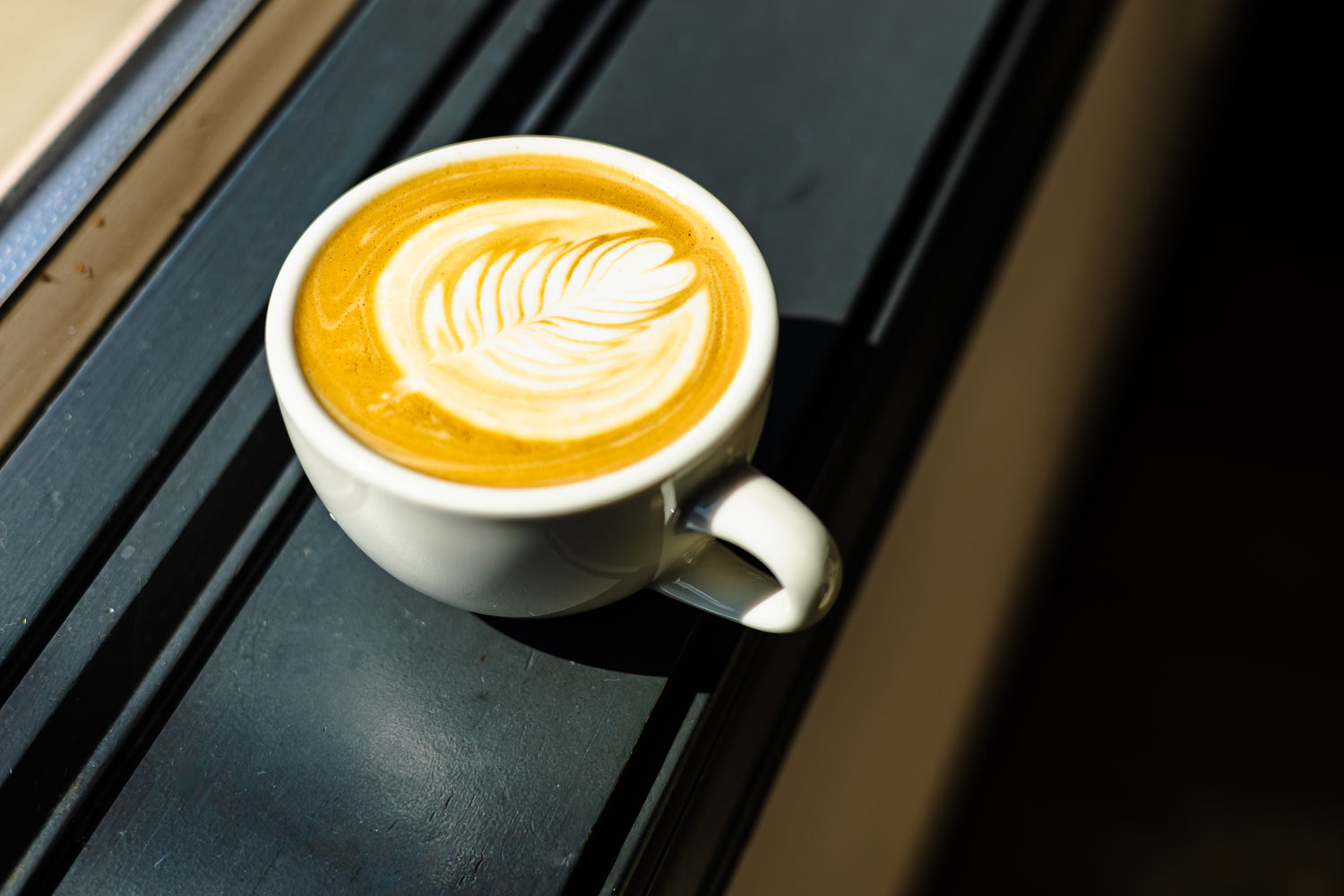 A white ceramic cup filled with a latte or cappuccino, featuring a leaf-shaped latte art design on top, resting on a dark wooden surface.