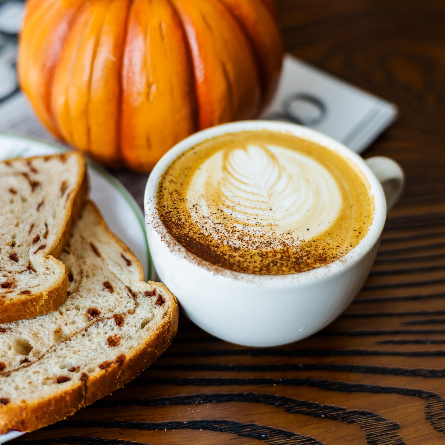 A white ceramic cup of latte coffee with leaf art foam and cinnamon dusting, placed on a dark wooden table next to slices of bread and a small orange pumpkin.
