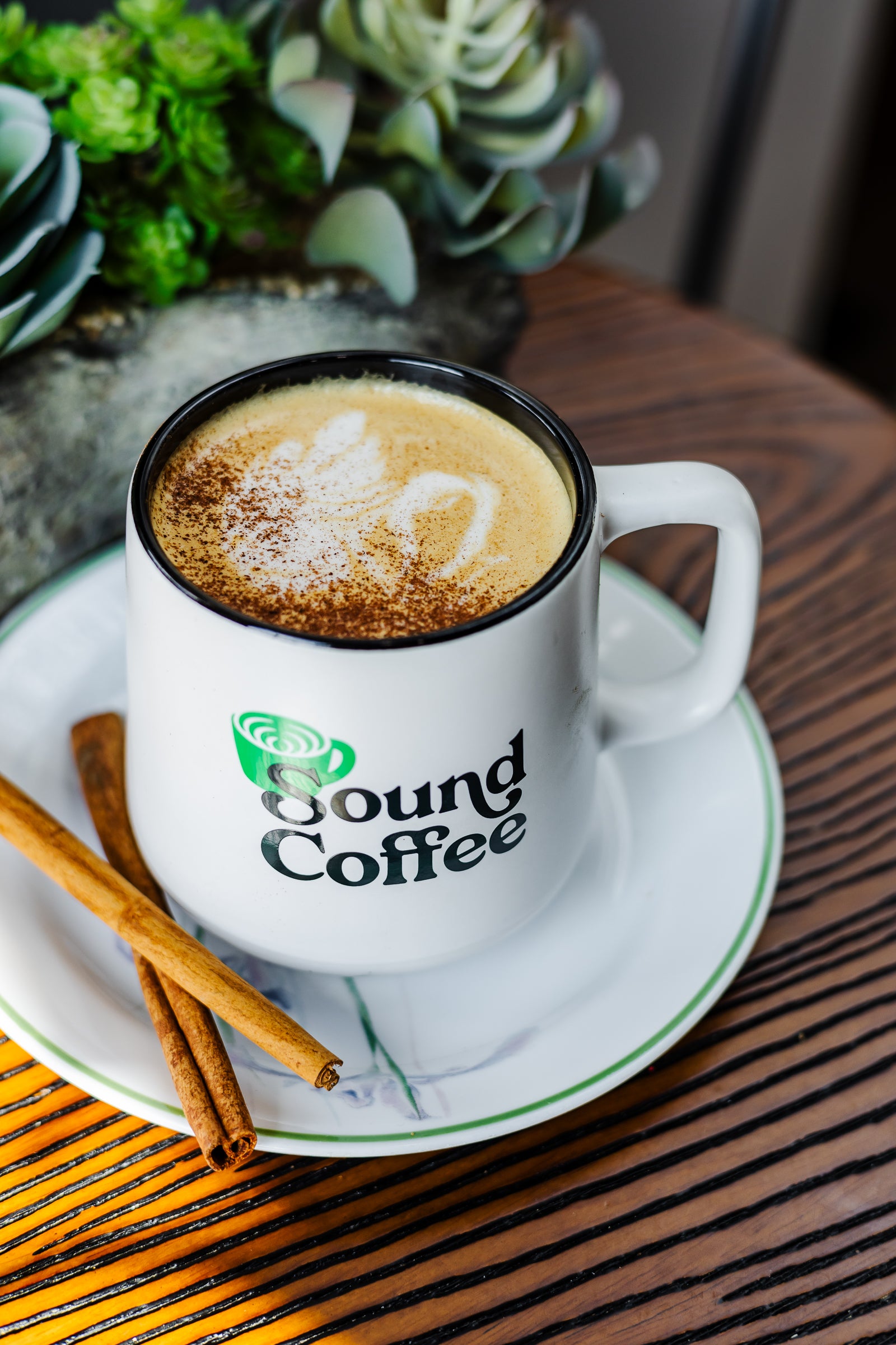 A white ceramic coffee mug with the 'Sound Coffee' logo, filled with a cappuccino topped with foam and cinnamon, sitting on a matching saucer with cinnamon sticks on a wooden table.