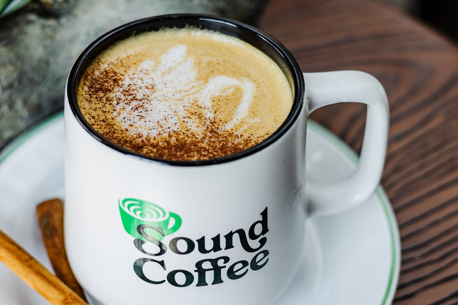 A white ceramic coffee mug with the 'Sound Coffee' logo, filled with a cappuccino topped with foam and cinnamon, sitting on a matching saucer with cinnamon sticks on a wooden table.