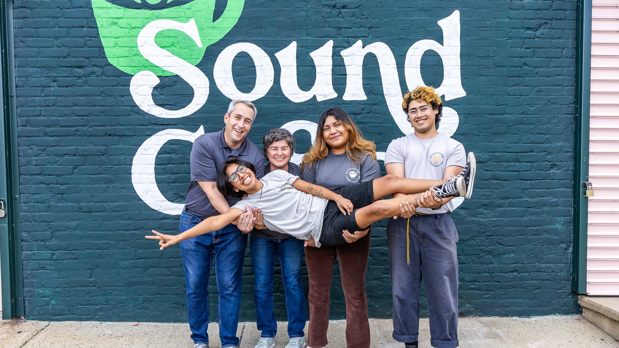 Five people posing together in front of a dark teal brick wall with a large white 'Sound.