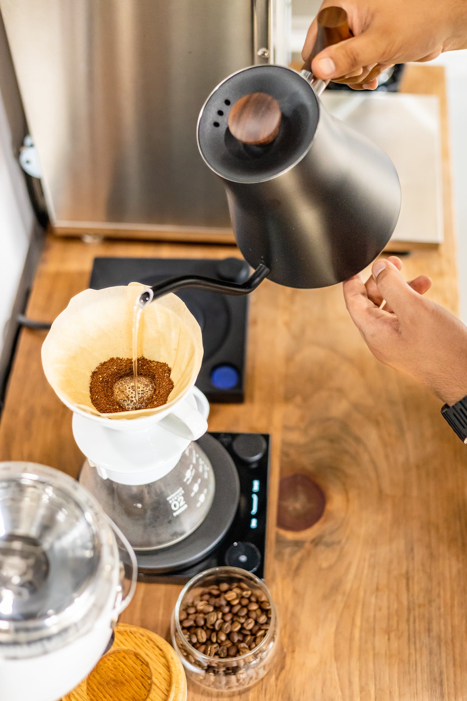 A close-up of hands pouring hot water from a matte black gooseneck kettle into a white paper cone filter holding coffee grounds over a glass carafe, set on a digital scale on a wooden countertop.
