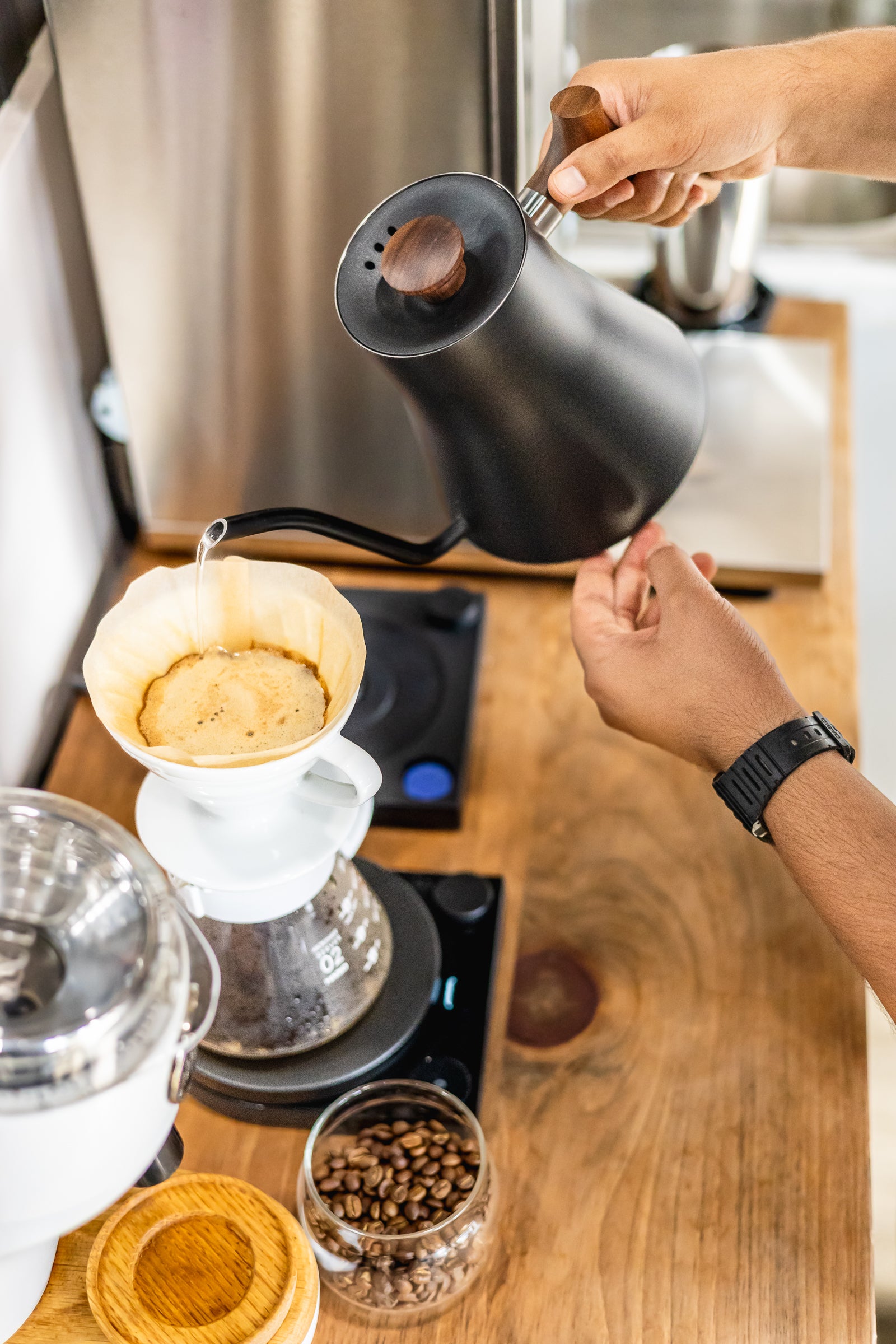 A person pouring hot water from a matte black gooseneck kettle into a white pour-over coffee dripper on a glass carafe, set on a digital scale over a wooden countertop with coffee beans nearby.