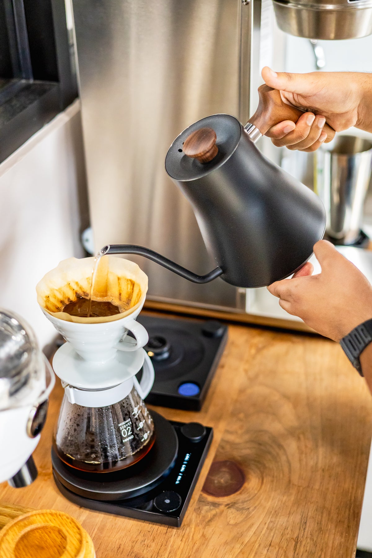A person pouring hot water from a matte black pour-over kettle into a white cone filter on a glass coffee dripper over a carafe, set on a digital burner on a wooden countertop.