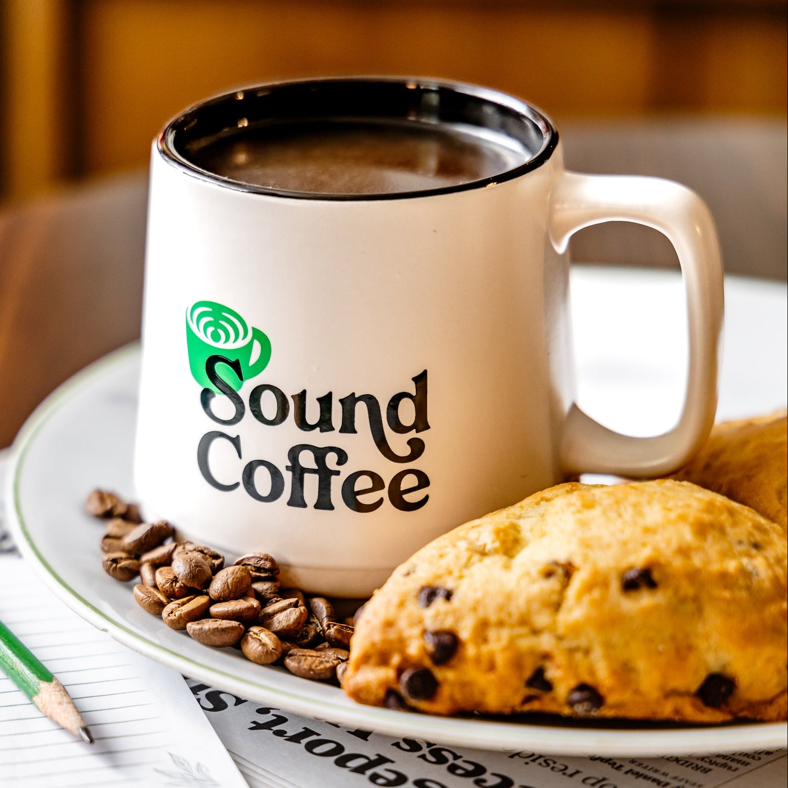 A white ceramic coffee mug with a black rim and handle, printed with the green and black 'Sound Coffee' logo, filled with coffee and styled on a plate with coffee beans, a chocolate chip scone, and newspaper.
