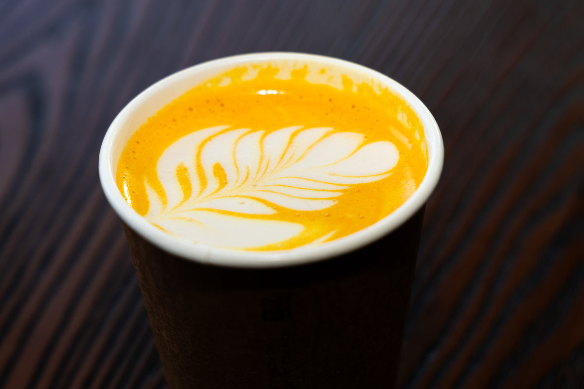 A close-up of a takeaway coffee cup filled with an orange-yellow drink and white leaf latte art, set on a dark wood table.