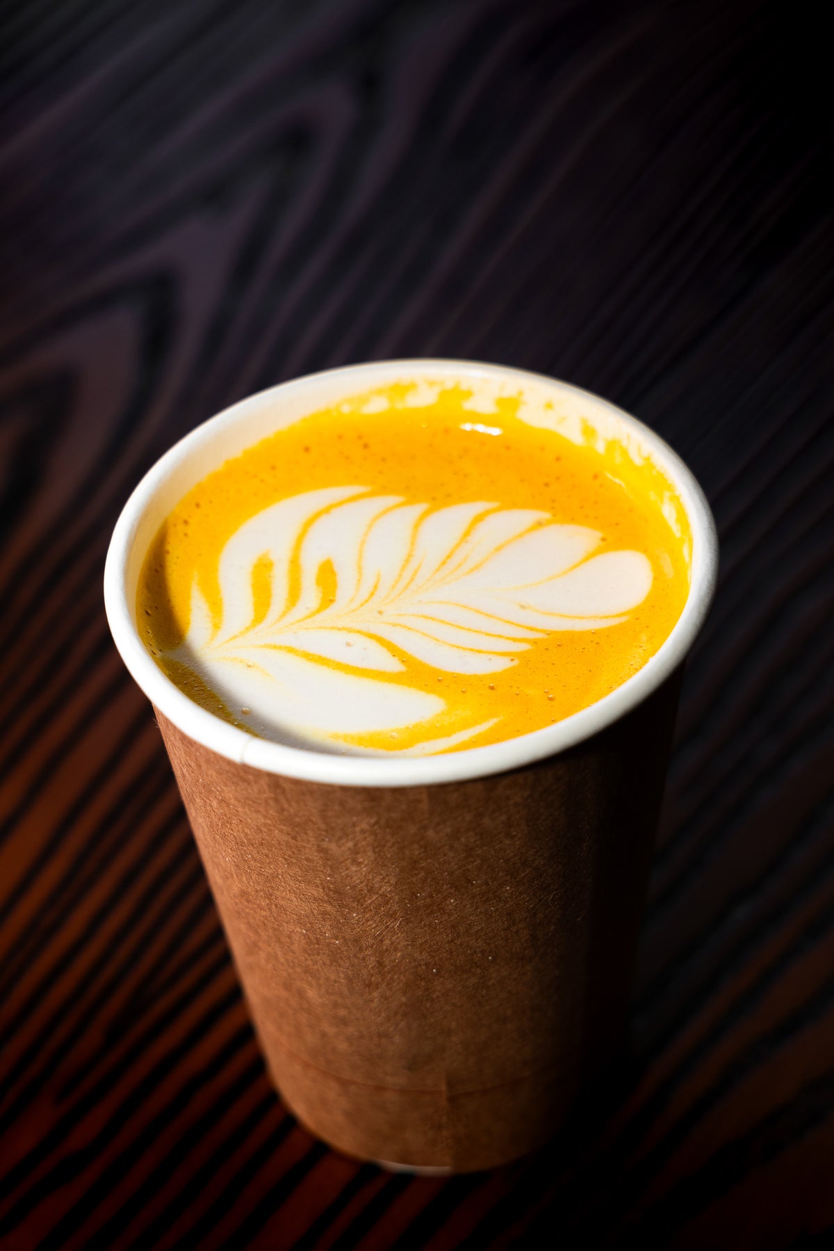 A paper coffee cup filled with a warm orange-yellow drink topped with white latte art, set on a dark wood surface.
