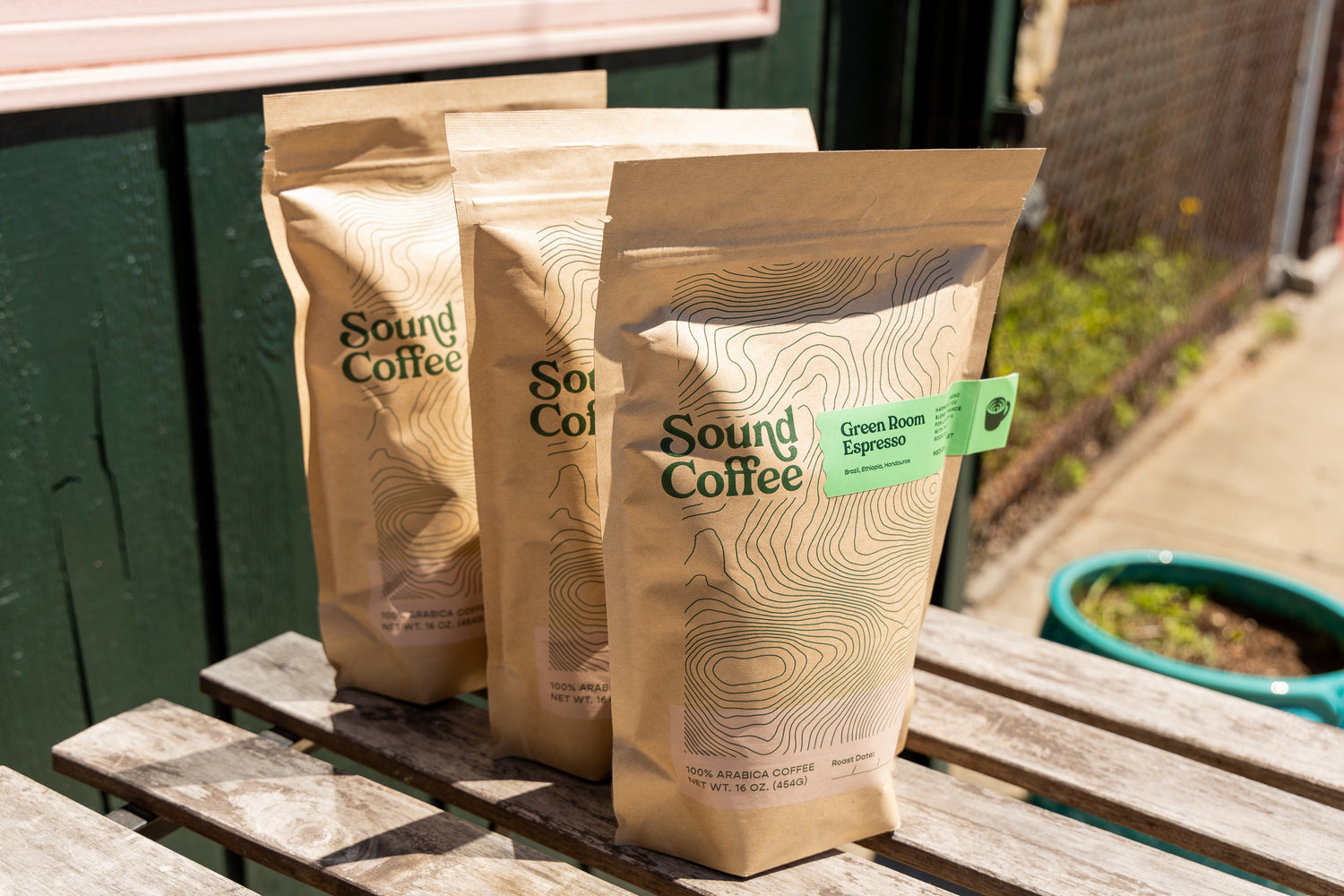 Three bags of Sound Coffee Green Roast Espresso standing on a weathered wooden table outdoors, with a green fence and garden planter in the background.