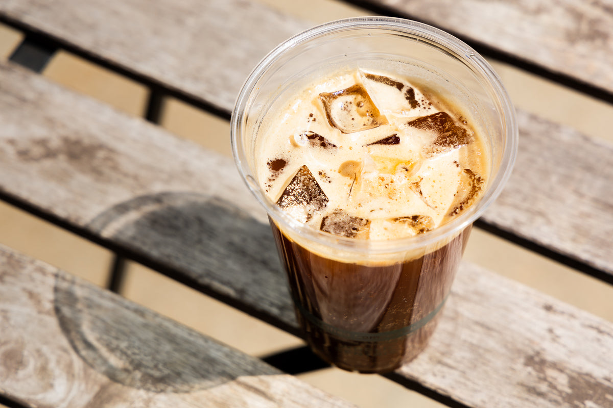 A clear plastic cup filled with iced coffee and ice cubes, placed on a weathered wooden slat surface.