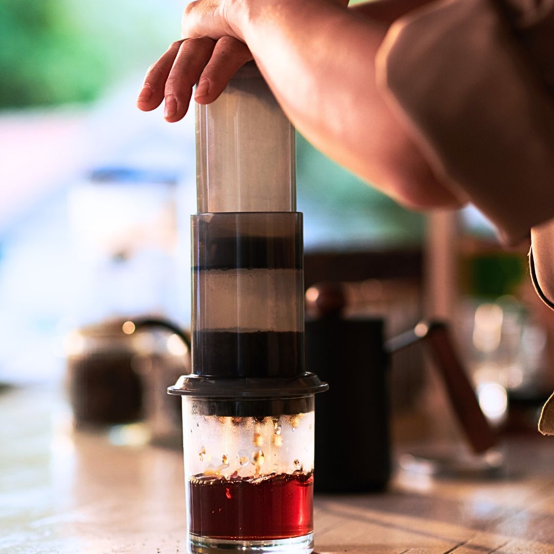 A close-up lifestyle shot of a person's hand operating a manual pour-over coffee maker, with dark coffee dripping into a glass carafe on a wooden table.