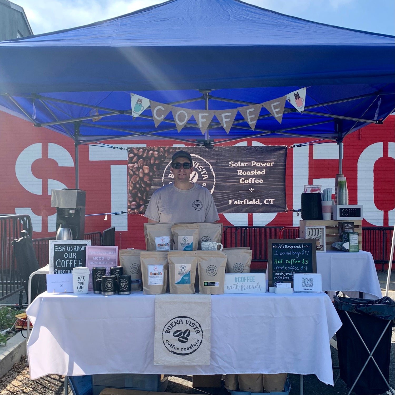 A coffee booth under a blue canopy tent, featuring a person behind a table with coffee bags, mugs, and small signs. The booth has a white tablecloth, and a banner reading 'COFFEE' hanging above.