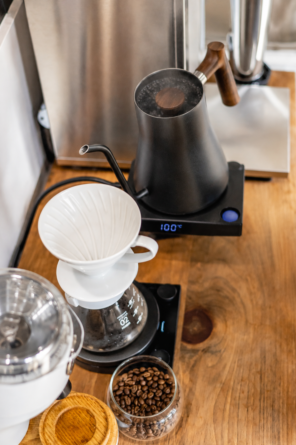 A pour-over coffee setup on a wooden countertop, featuring a white ceramic dripper over a glass carafe, a black gooseneck kettle on a digital scale, and a small glass bowl of coffee beans.