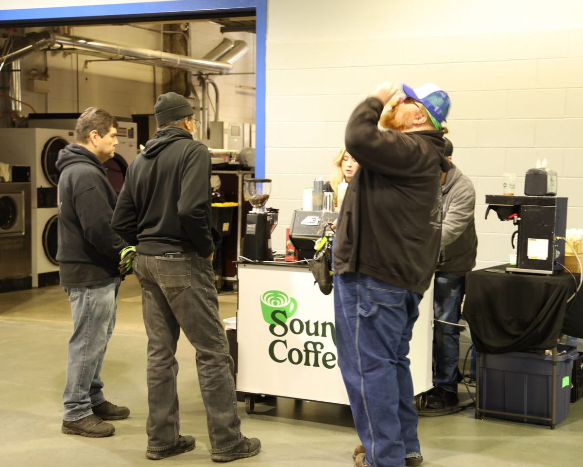 A group of people gathered around a coffee cart in a workshop or warehouse setting. The cart has a white sign that reads 'Sound Coffee' with a green coffee cup logo.