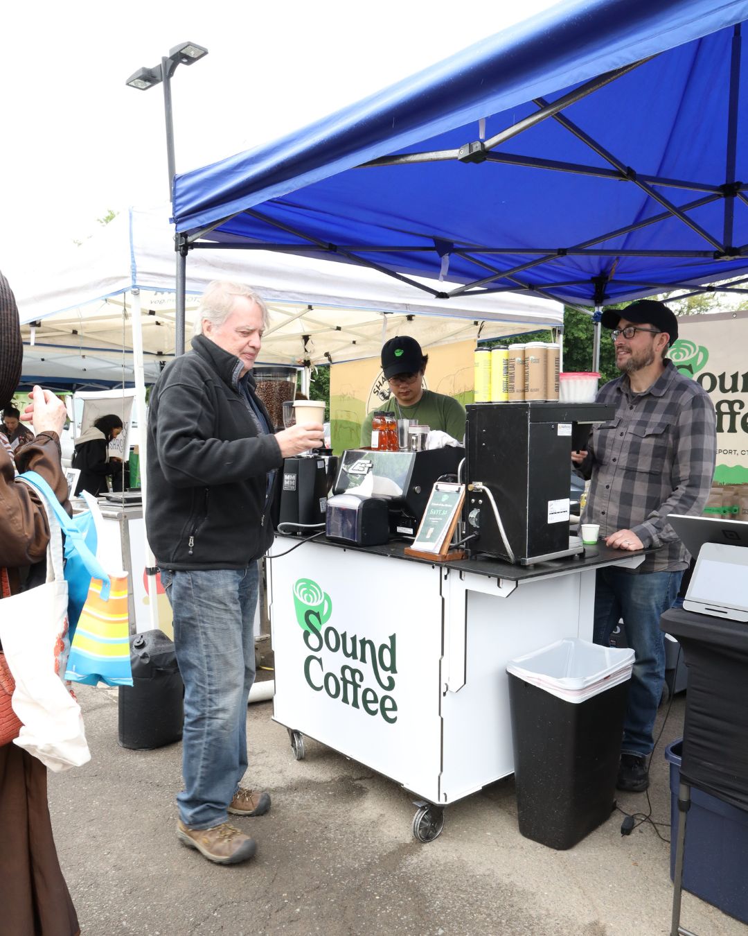 A man orders coffee from a mobile coffee station at an outdoor market. The white cart branded 'Sound Coffee' is under a blue canopy, with a vendor behind it and another man standing nearby.