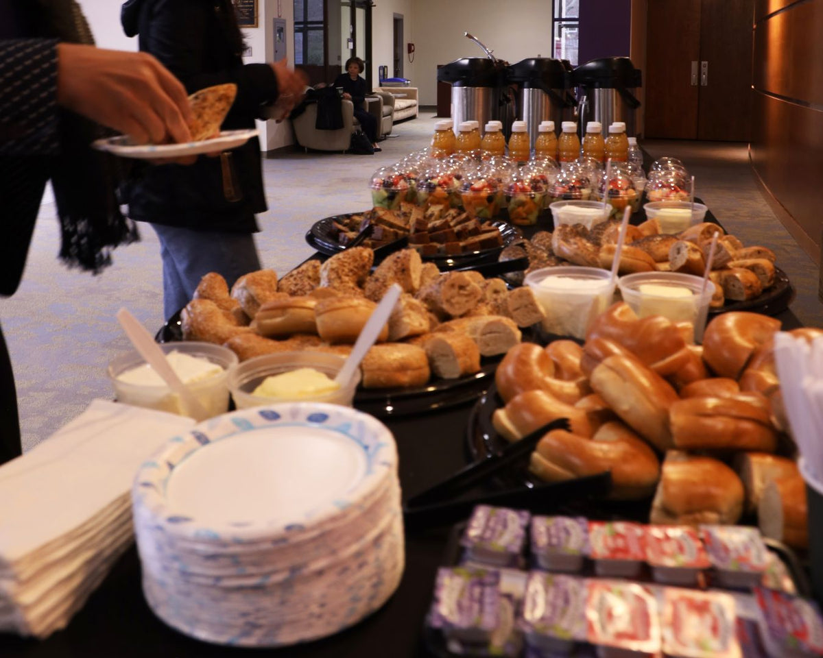 A buffet-style food table in a lobby or cafeteria setting, filled with assorted bagels, pretzels, bread rolls, small salads, cups of dip, bottled drinks, and stacks of paper napkins. A person's arm and torso are visible on the left holding a plate.