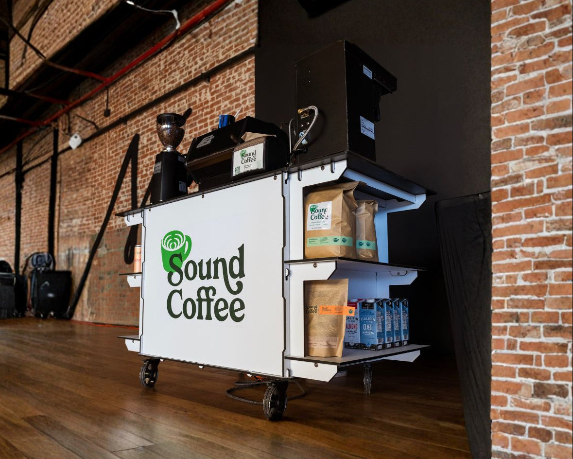 A white Sound Coffee cart with shelves and a top bar, shown in a loft-style room with brick walls and wood floors. The cart has the Sound Coffee logo on the side and is stocked with coffee bags and bottled drinks.