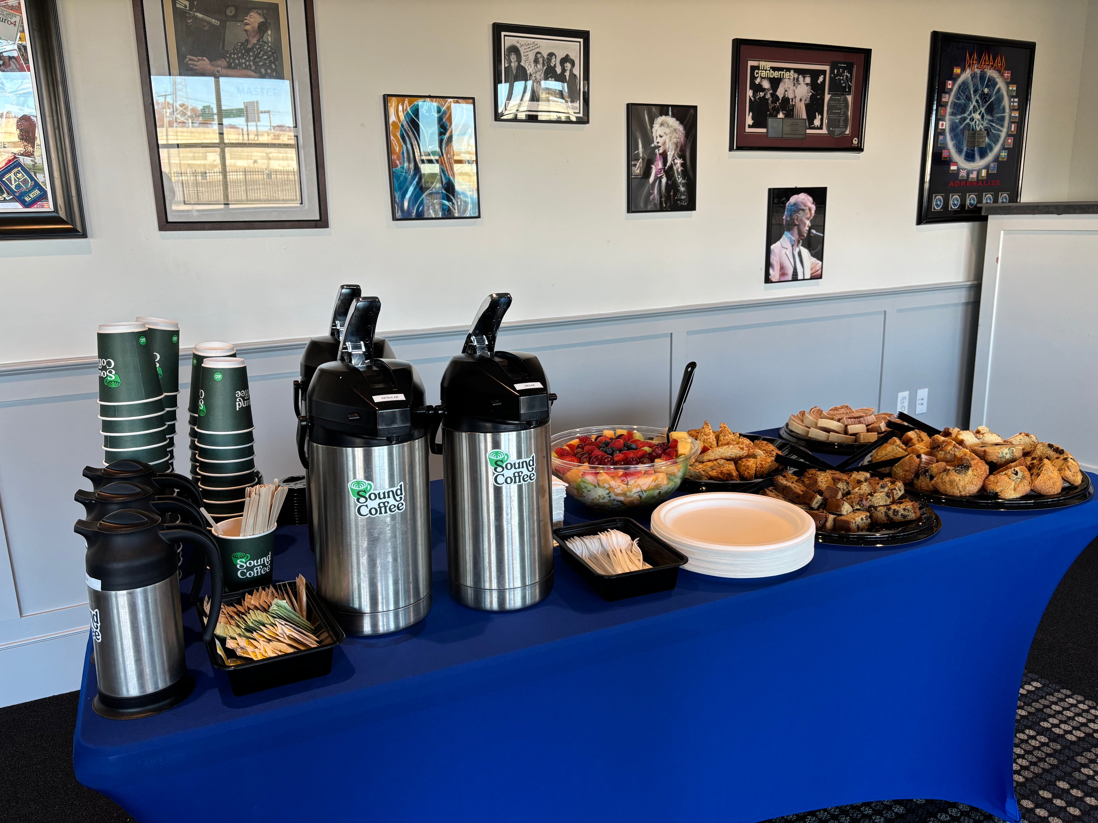 A catering buffet table covered with a bright blue tablecloth, stocked with coffee thermos pitchers, paper cups, fruit, pastries, and baked goods, set against a wall with framed pictures.