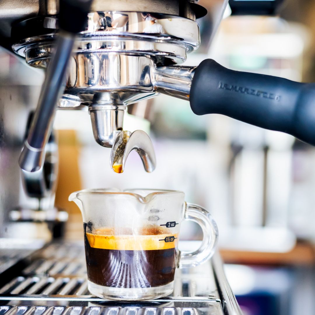 A close-up of espresso dripping from a metal portafilter with a black Marzotto handle into a small clear glass measuring cup filled with dark coffee.