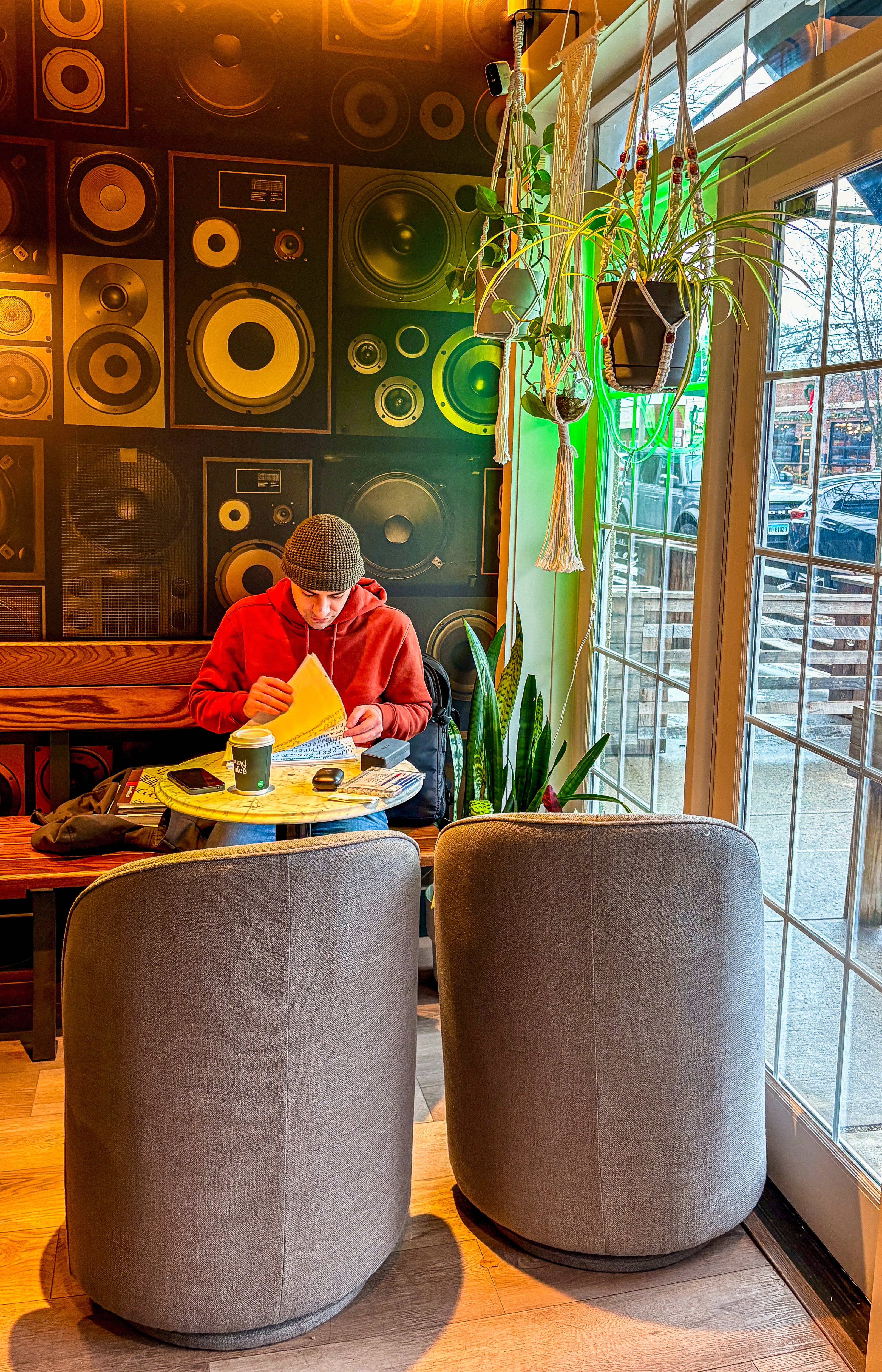A cozy cafe interior with a person in a red hoodie and beanie sitting at a small table, surrounded by two large gray upholstered chairs, plants, and a wall covered in circular speaker designs. Large windows let in warm light, and a street scene is visible outside.