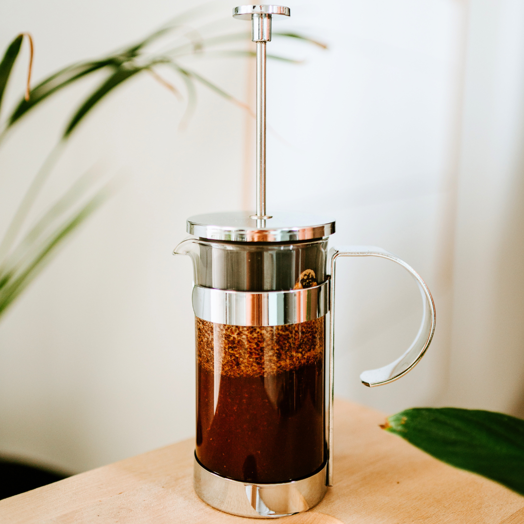 A stainless steel and glass French press coffee maker filled with dark coffee sits on a light wooden table, with blurred green leaves in the background.