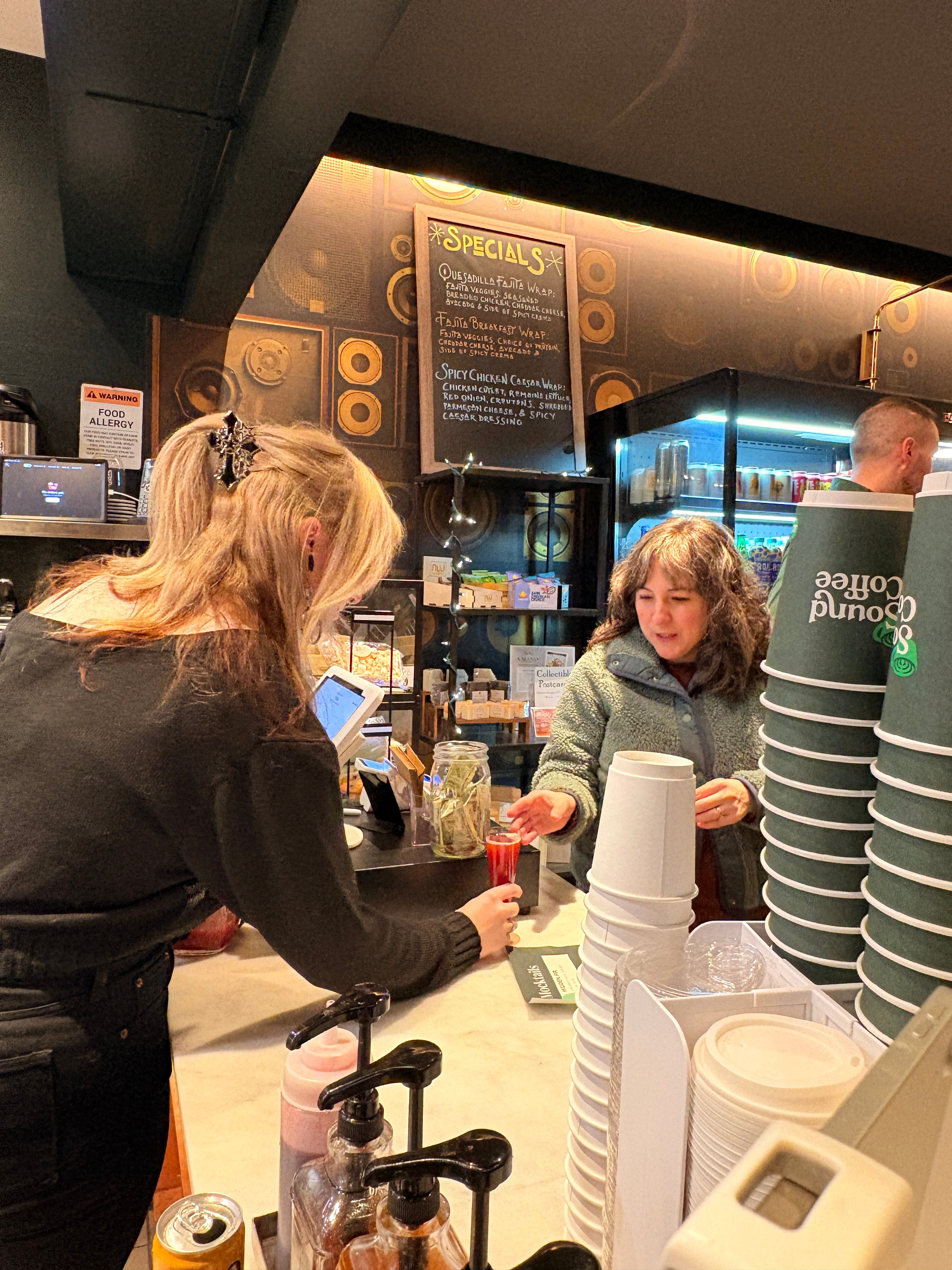 A busy coffee shop counter scene with stacks of white cups, a person buying a drink, and another person preparing a drink behind the counter.