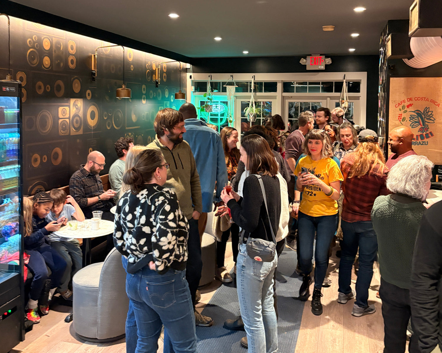 A crowded indoor event or gathering in a modern space with wood flooring, black walls, and multiple people standing and sitting. No single retail product is clearly isolated as the subject.