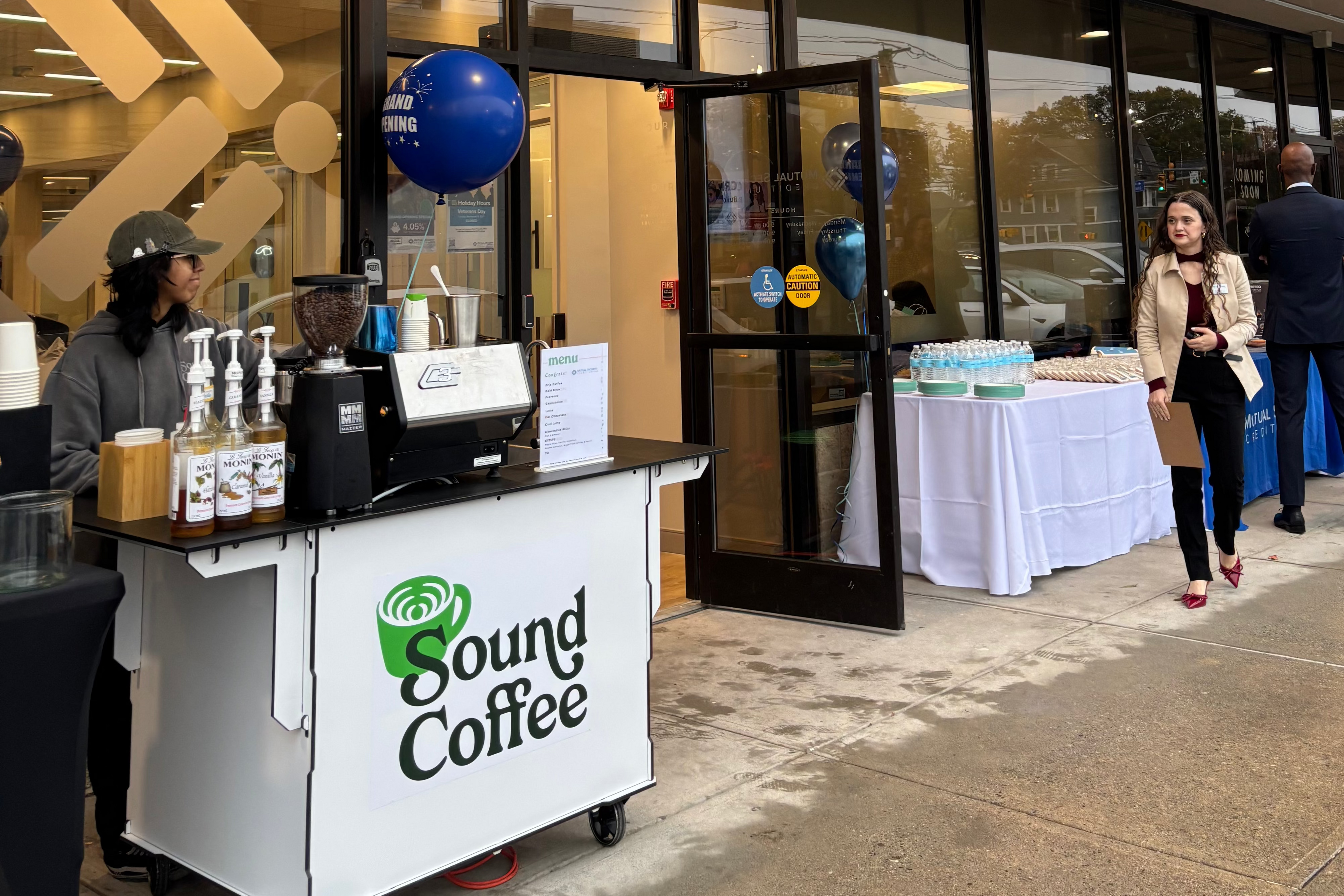 A storefront scene with a Sound Coffee cart set up outside on the sidewalk, with several people standing near the entrance and a building facade above with large signage.