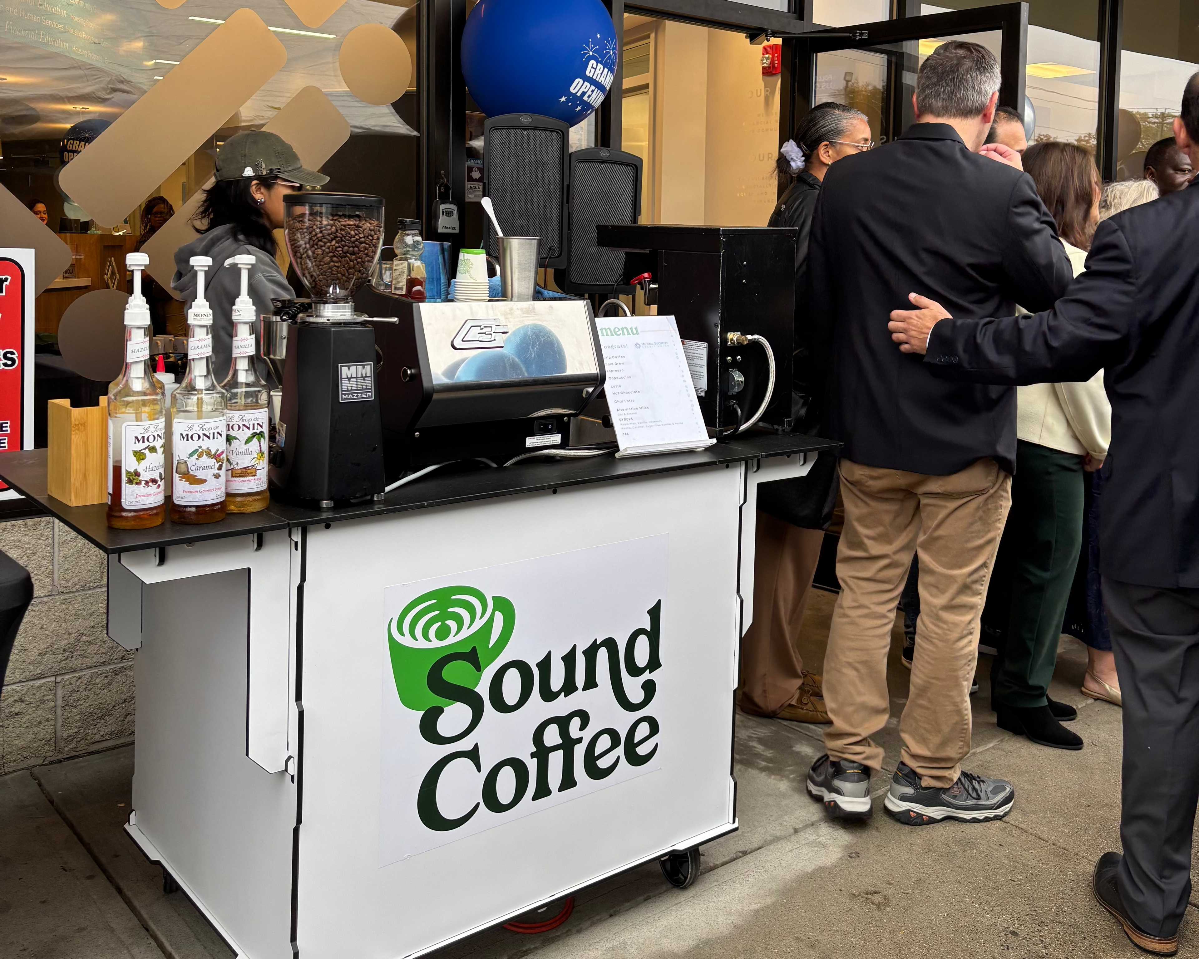 A white Sound Coffee cart with a green logo stands on a sidewalk in front of a storefront, with several people gathered around it.