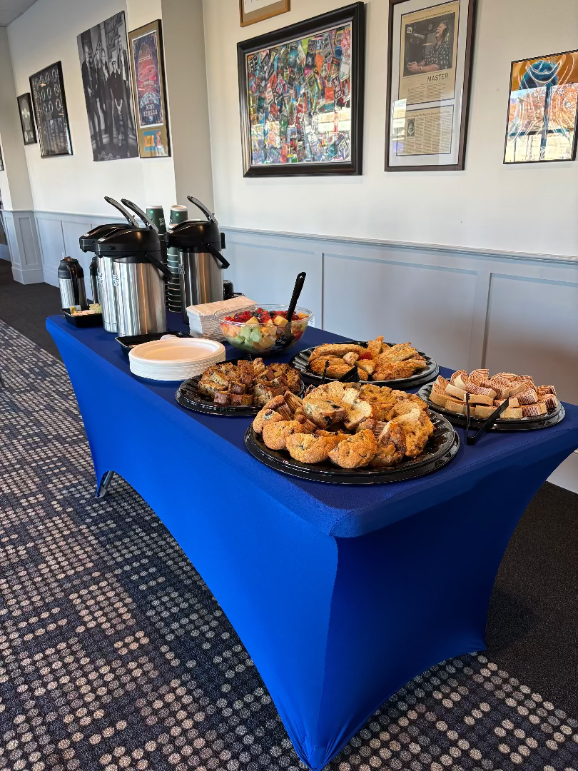 A catering buffet table covered in a bright blue tablecloth, filled with assorted pastries, breads, and salads, set in an indoor event space with framed wall art and carpeted floor.