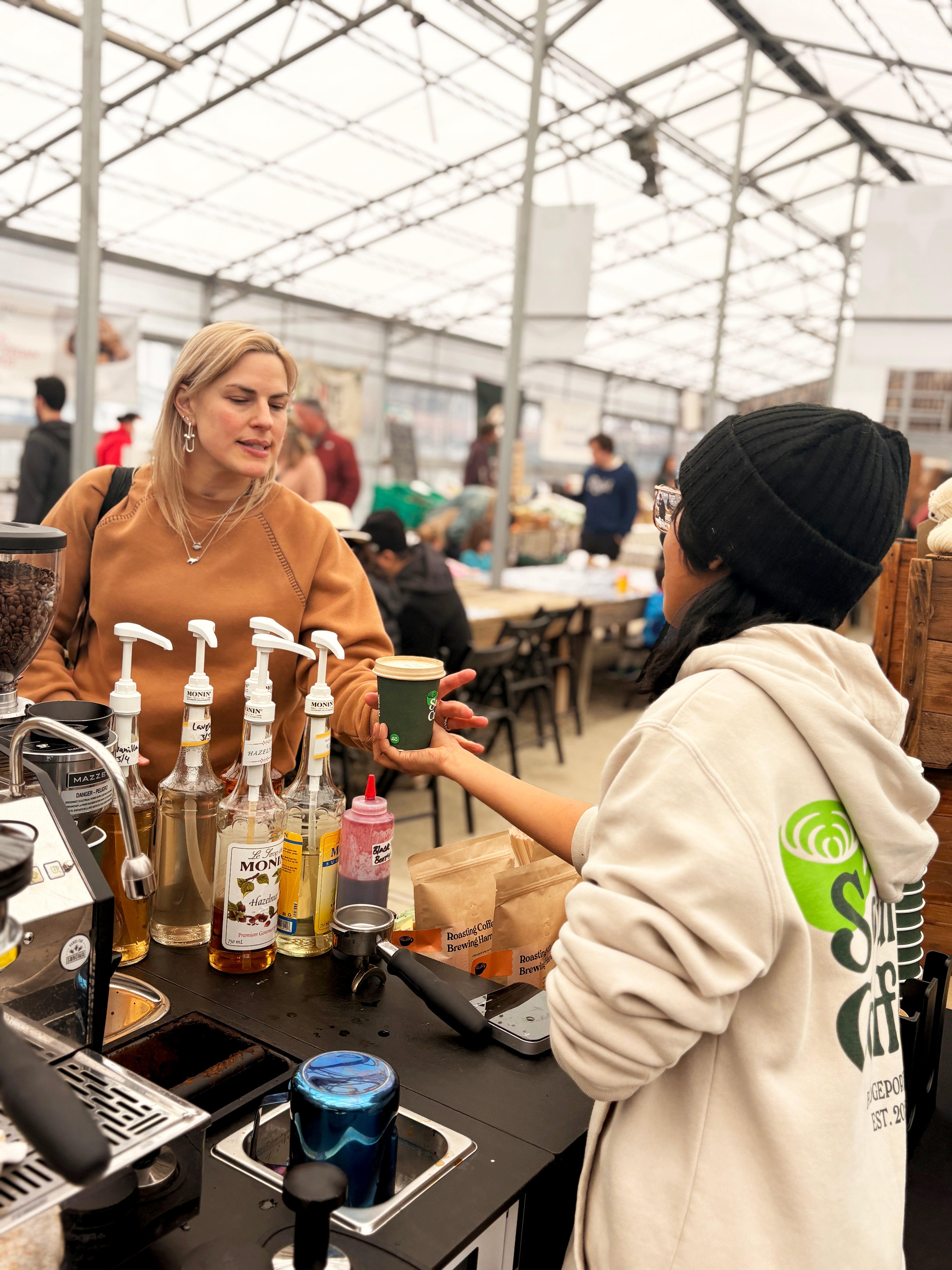 A person in a black beanie and cream hoodie gives a green paper coffee cup to a woman in a tan sweater at an indoor cafe counter with syrup bottles and espresso equipment.