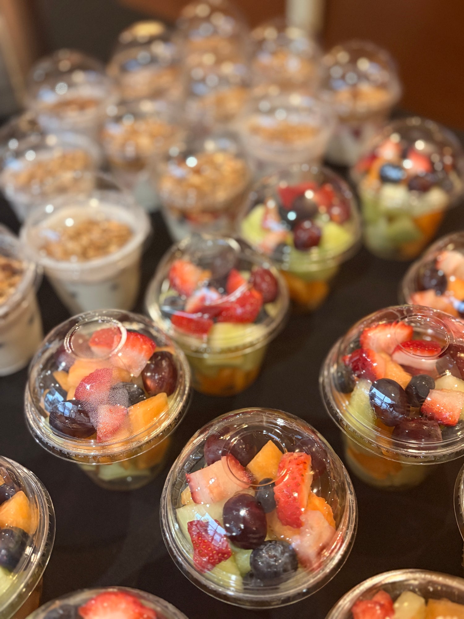 Multiple clear plastic cups filled with fruit parfaits, including strawberries, blueberries, grapes, and granola, arranged on a dark table.