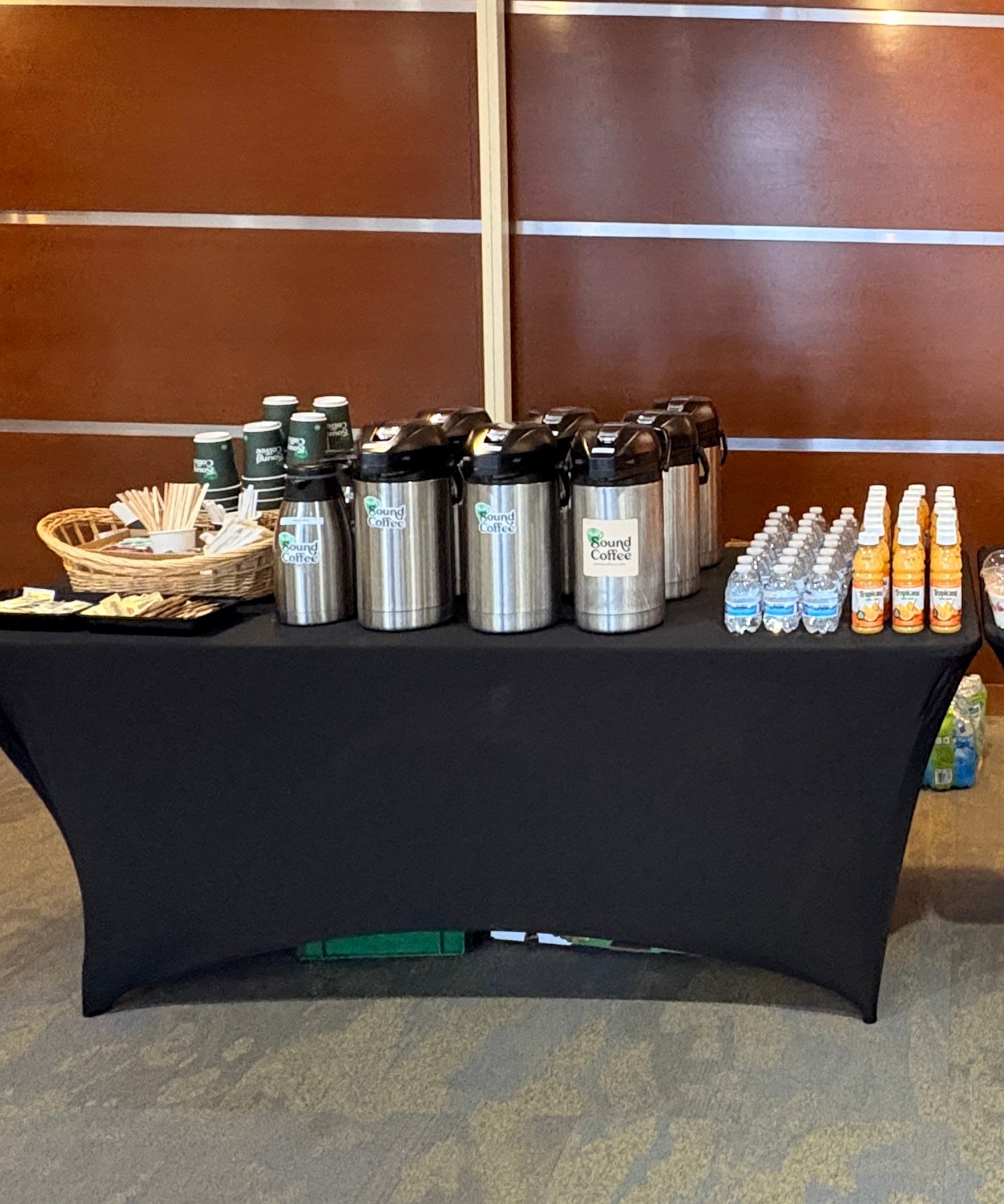 A beverage station on a black draped tablecloth with stainless steel coffee urns, bottled drinks, and a basket of utensils in front of a brown wood-paneled wall.