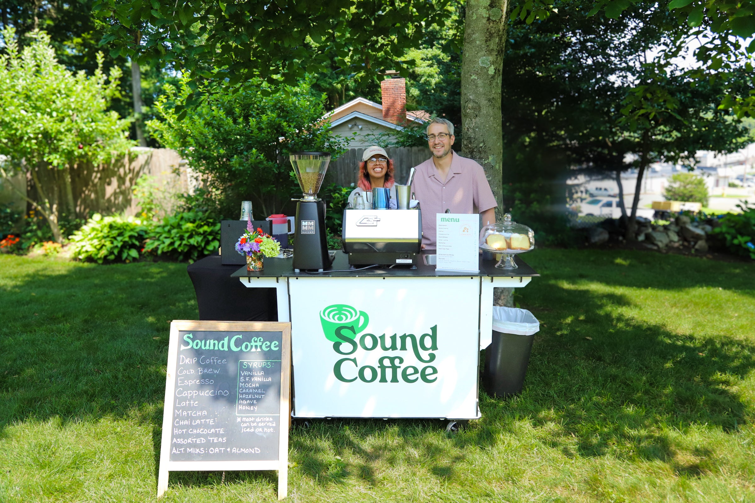 A small outdoor coffee cart with two people standing behind it on a grassy lawn. The cart has a white front panel with the green 'Sound Coffee' logo, coffee equipment, a sign board, and a chalkboard menu sign.