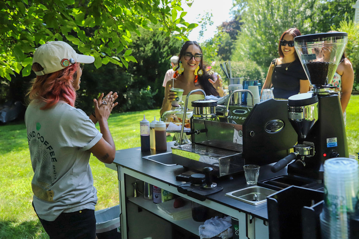 Outdoor coffee station scene with people gathered around an espresso machine and coffee grinders on a counter, set in a grassy park area with trees in the background.