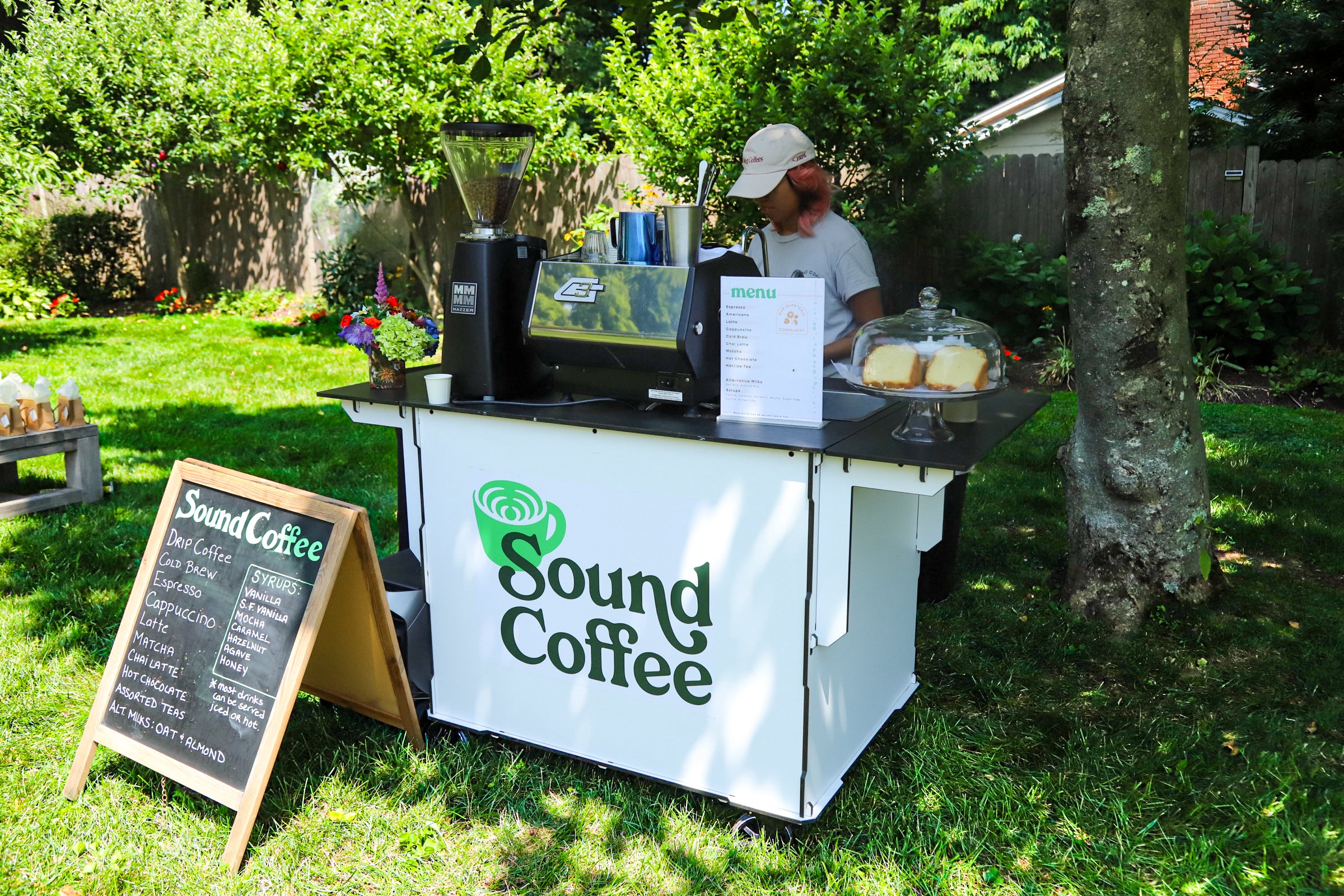 A white Sound Coffee cart set up outdoors on a grassy lawn next to a tree, with a person standing behind it. A black espresso machine, coffee grinder, and a chalkboard sign listing menu items are visible on the cart and on the ground.
