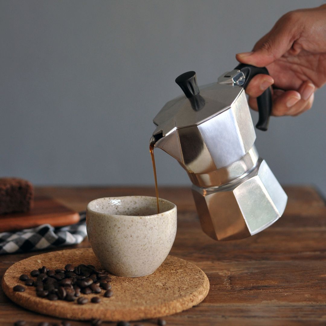 A hand holds a silver aluminum moka pot and pours coffee into a speckled beige ceramic cup on a cork coaster, with coffee beans scattered nearby on a wooden table.