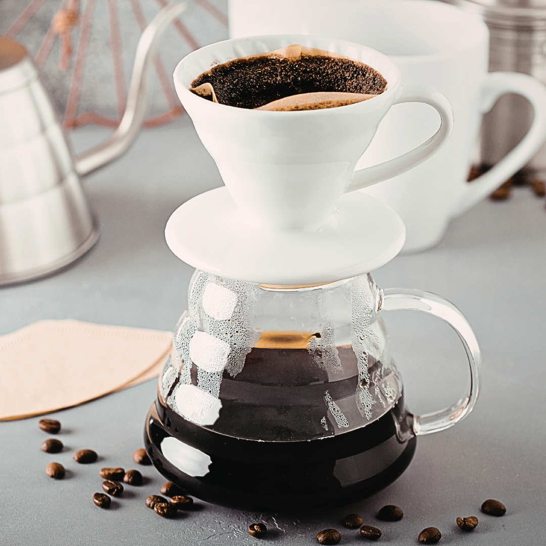 A pour-over coffee setup with a white ceramic dripper filled with coffee above a clear glass carafe containing brewed black coffee, styled on a gray surface with scattered coffee beans and a metal kettle in the background.