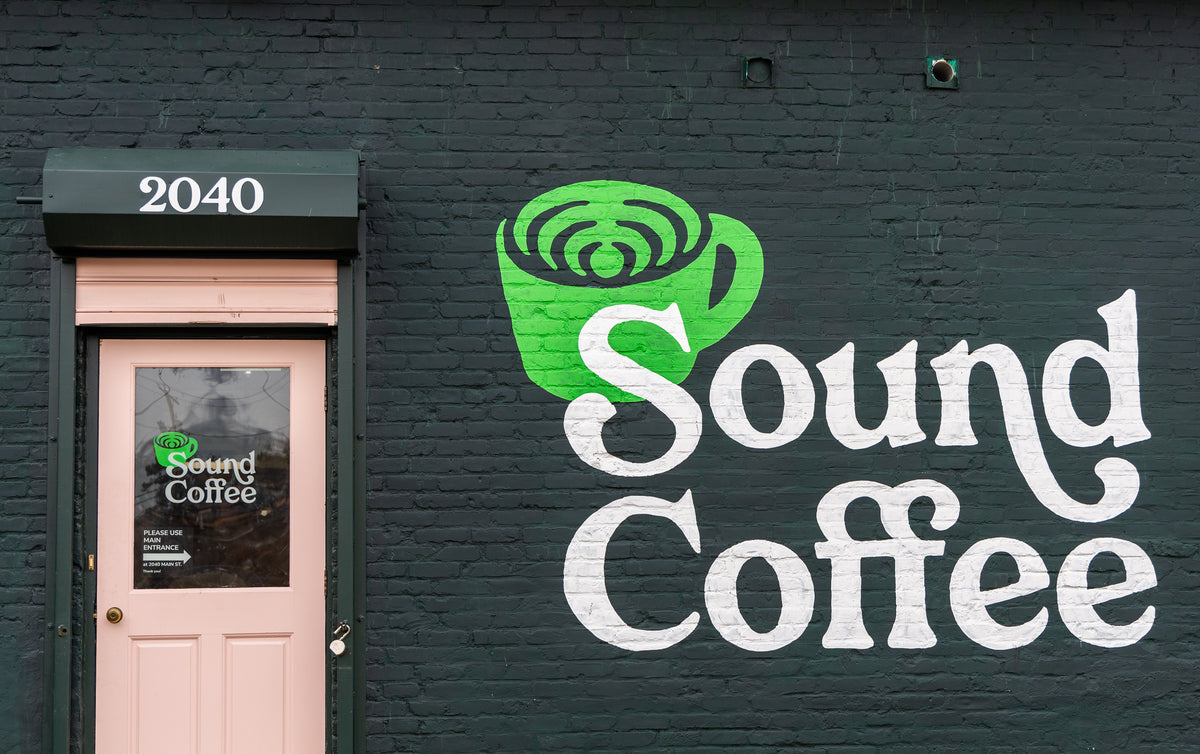 A storefront wall painted dark green with a large white 'Sound Coffee' logo and a green coffee cup graphic. A pale pink door with the number 2040 is visible on the left.