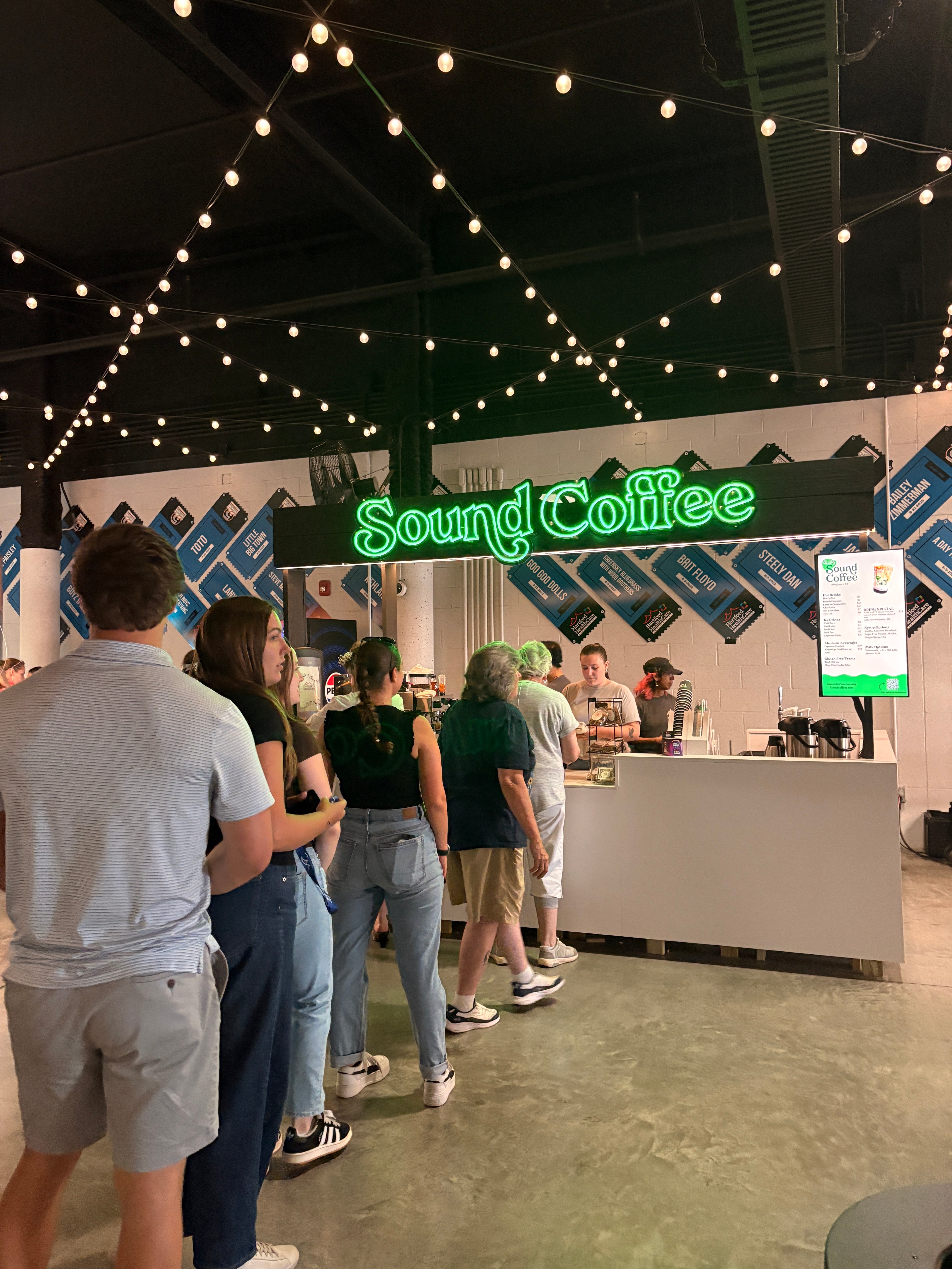 A group of people standing in a line at a Sound Coffee kiosk counter at the Amp, with a bright green neon sign and string lights visible in the background.